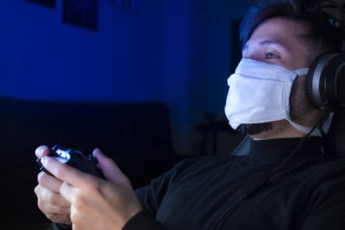 Adam ameliyat maskesi takıp kulaklıkla video oyunu oynuyor. Kavram: Coronavirüs için kendini izolasyon (COVID-19) Video oyunları oynama zamanı.
