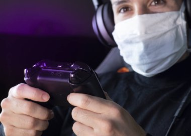 Covid-19 karantinaya alınırken genç beyaz adam yüzünde tıbbi maskeyle video oyunları oynuyor.