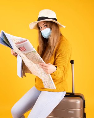 Tıbbi maskeli bir turist kız, bavullu, şapka haritaya bakıyor, uçuşlar covid-19 tarafından iptal edildi.