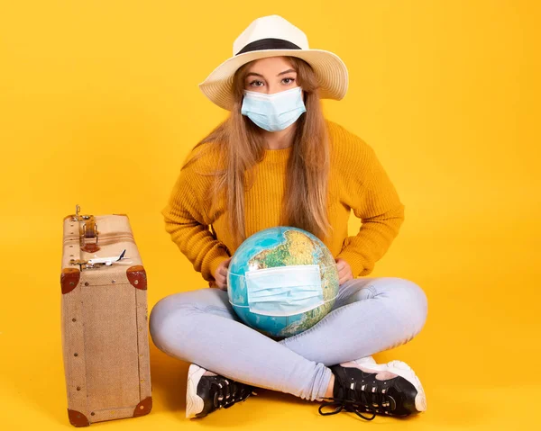 A tourist girl with a medical mask, has a suitcase, a globe, is willing to travel but the covid-19 prevents it