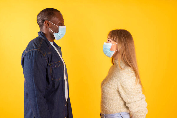 couple look at each other with medical mask, interracial profile view, on yellow background