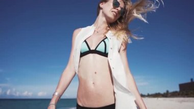 Kız, plaj, deniz, saç A güzel girl renkli bikini ve şeffaf beyaz pelerini rüzgarda kum dizlerinin üzerine oturur ve ince kumlu plaja elleriyle yükseltir ve dökülen