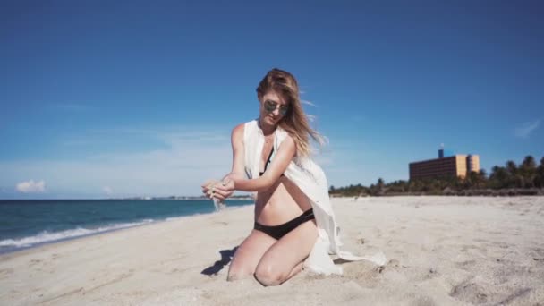 Meisje, strand, zee, wind in je haren A beautiful girl in een gekleurde bikini en een transparante witte mantel, zit op het zand op haar knieën en verhoogt haar handen met fijn zand van het strand en giet het — Stockvideo