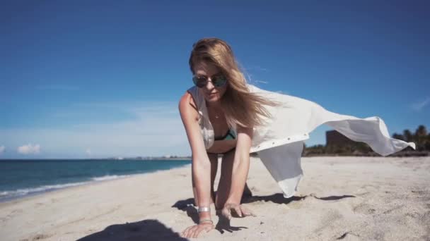 Дівчина, пляж, море, вітер у волоссі Красива дівчина в кольоровому бікіні і прозорий білий плащ, сидить на піску на колінах і піднімає руки з дрібним піском з пляжу і виливає його — стокове відео
