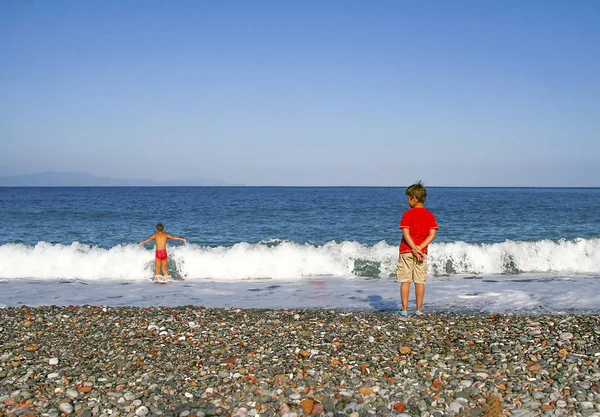 Two boys , one standing and looking on the beach and the other playing with big waves.