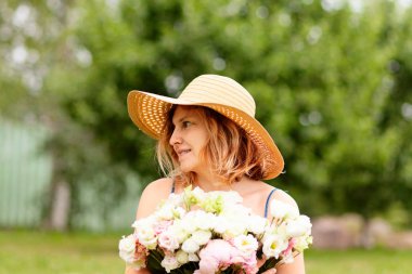 Genç bir kadın büyük bir buket şakayık tutuyor. Geniş kenarlı hasır bir şapkayla. Sağa bakıyor..