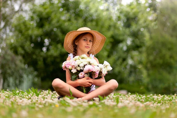 女の子牡丹の大きな花束を保持します。若いですティーンでA帽子で広いつばは、開花草原に座っています. — ストック写真