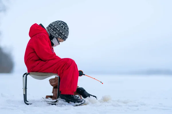 Um rapaz de fato vermelho num lago gelado de neve. Vara de pesca nas mãos de um menino. Pesca de inverno no buraco de gelo . — Fotografia de Stock