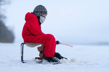 Kış balıkçılığında kırmızı tulumlu küçük bir çocuk. Katlanan bir sandalyede oturuyor. Bir oltanın ellerinde.