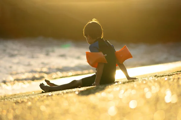 Ein Junge in Rüschen und Neoprenanzug sitzt am Strand. Das Kind blickt auf das Meer. — Stockfoto
