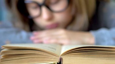 Ön planda bir kitap var. Büyük siyah gözlüklü bir kız kitap okuyor. Görüntüyü kapat.