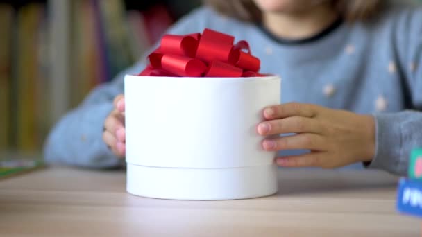 Mooie witte doos met een rode strik in de handen van een kind. Een klein meisje opent een geschenkdoos met een verrassing. Close-up zicht. — Stockvideo