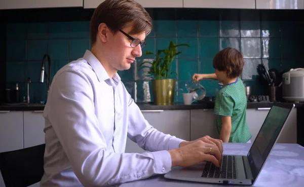 Молодий чоловік працює на кухні в ноутбуці. Батько з сином. Домашній офіс з дітьми . — стокове фото