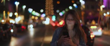Caddenin ortasında telefonuyla müzik dinleyen genç bir kadın. Arka planda Bokeh ışıkları var. Sığ dof, gerçek zamanlı, Bmpcc 4k