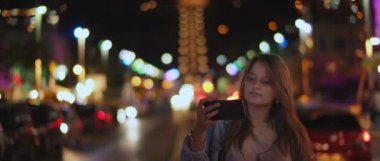 Sokağın ortasında selfie çeken genç bir kadın. Gece Bokeh ışıkları. Sığ dof, gerçek zamanlı, Bmpcc 4k