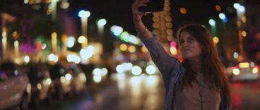 Caddenin ortasında selfie çeken ve gülümseyen genç bir kadın. Gece lambaları. Sığ dof, gerçek zamanlı, Bmpcc 4k