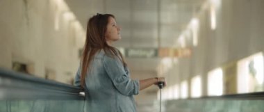 Rüzgar estiren uzun kahverengi saçlı ve kot gömlekli bir kadın yürüyen merdivene yaslanmış, elinde bavuluyla yukarı bakıyor. Arkadan çekim, ağır çekim, Bmpcc 4k