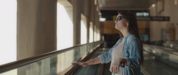 提着手提箱 太阳镜和斜纹棉布衬衫的女人站在机场的走道上 看着外面 脸上闪烁着光芒 慢动作 电影镜头 Bmpcc — 图库视频影像