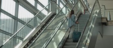 Uzun saçlı, kot gömlekli ve kot pantolonlu bir kadın havaalanında elinde turkuaz bavuluyla yürüyen merdivenle yukarı çıkıyor. Ağır çekimde, Bmpcc 4k.