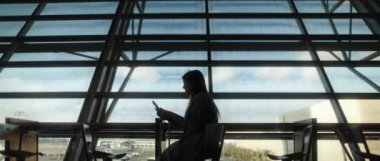 Büyük pencerenin önünde oturan ve havaalanı terminalinde uçağını beklerken telefonunu kaydıran bir kadın. Yavaş çekim, Bmpcc 4k