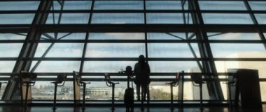 Terminal penceresinin önünde oturan siluet kadın, uçuşunu bekliyor. Boş salon, havaalanı salonundaki koltuklar. Büyük pencerenin dışındaki bulutlu gökyüzü