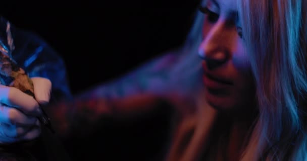 Gros plan d'une tatoueuse dessinant un tatouage sur la peau d'une personne. Mouvement lent, éclairage studio bleu, faible profondeur de champ. BMPCC 4K 