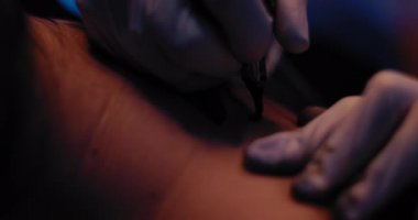 Dövme sanatçısının elinin, birinin sırtına dövme çizimi yaparken yakın plan. Yavaş çekim, mavi stüdyo ışıkları, sığ alan derinliği. BMPCC 4K