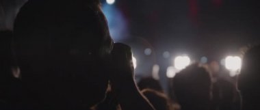 A man taking a picture of band performance during live show concert, while people crowd jumping and dancing in front of a stage of a night club. Medium, behind shot, handheld, slow motion. BMPCC 4K