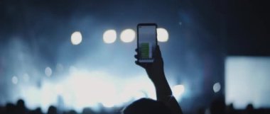Person taking a picture during a concert, with floodlights flashing in the background. People in front of a stage of a live show at a night club. Medium, behind shot, handheld, slow motion. BMPCC 4K 