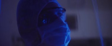 Koruyucu takım elbiseli, maskeli ve gözlüklü bir adamın etrafını kolaçan edin. Sağlık ve güvenlik kavramı, virüs koruması, salgın. Mavi ışık, yavaş çekim. BMPCC 4K