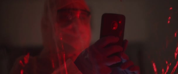マスクと眼鏡で保護スーツの男は Pvcシートの後ろに 携帯電話で写真を撮る ウイルス保護 パンデミックの概念 レッドライト スローモーション Bmpcc — ストック動画
