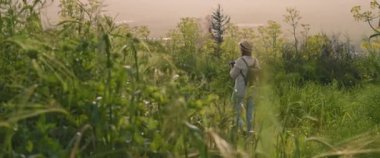 Sırt çantalı ve şapkalı genç bir kız yeşil alanda yürüyor, gün batımında manzara ve doğa fotoğrafları çekiyor. Yavaş hareket et, Shallow Dof. Macera, yaşam tarzı konsepti. BMPCC 4K 