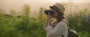 Sırt çantalı ve şapkalı genç bir kız güneş batarken manzara ve doğa fotoğrafları çekiyor. Yaklaş, yavaş hareket et, Shallow Dof. Macera, yaşam tarzı konsepti. BMPCC 4K 