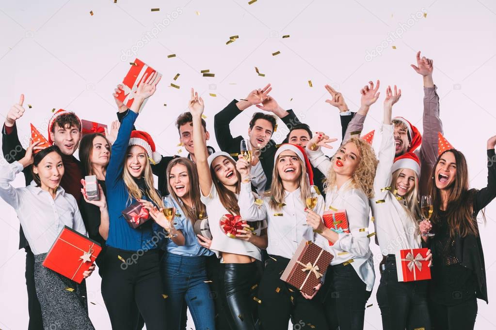 Party with friends. Group of cheerful young people in Santa hats with gift boxes showered with confetti.