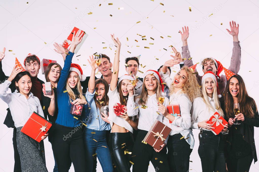Party with friends. Group of cheerful young people in Santa hats with gift boxes showered with confetti.