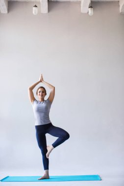 Pratik yoga, Vrksasana egzersiz namaste ile ayakta spor giyim kadın gülümsüyor, ağaç poz gri arka plan üzerinde.