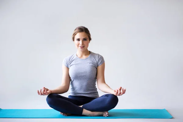 Hermosa joven en ropa deportiva practicando yoga mientras está sentada en posición de loto sobre fondo gris . —  Fotos de Stock