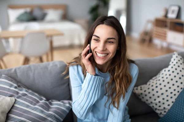 Hermosa mujer positiva hablando por teléfono mientras está sentada en un sofá en casa . —  Fotos de Stock