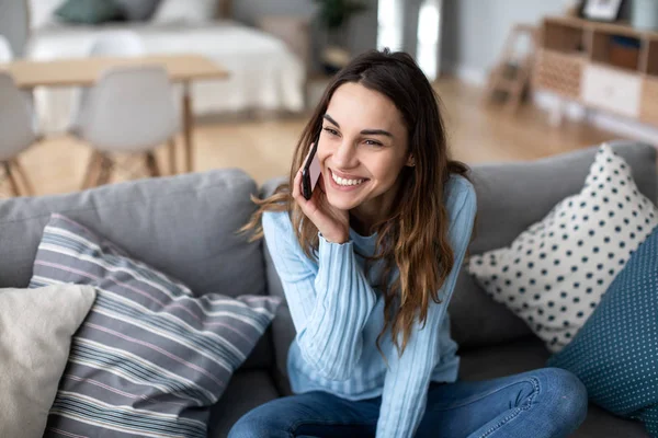 Atractiva joven mujer hablando por teléfono y sonriendo mientras está sentada en un sofá en casa . —  Fotos de Stock