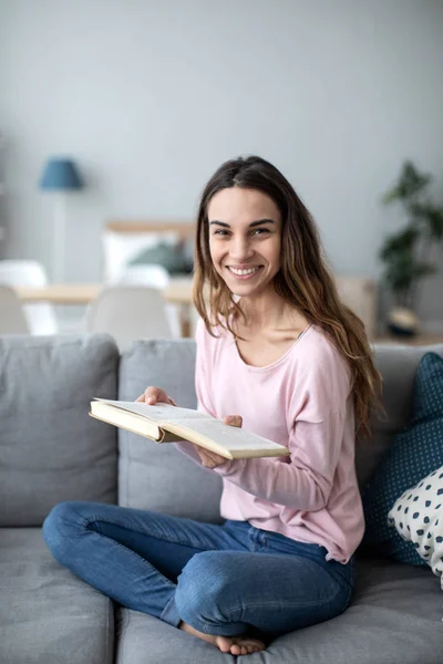 Mulher com um livro no sofá olhando para a câmera com um sorriso . — Fotografia de Stock
