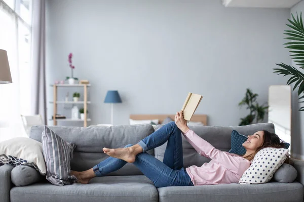 Joven hermosa mujer está leyendo un libro mientras está acostado en un sofá en la sala de estar . —  Fotos de Stock