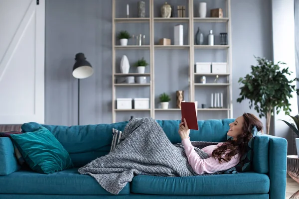 Mujer feliz con un libro en sus manos descansando en un sofá acogedor en h —  Fotos de Stock
