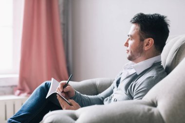 Psikolog. Koltukta oturan ve arka planda pembe perdelerle yazı yazan bir adamın yan fotoğrafı..