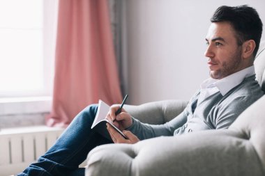 Psikolog. Koltukta oturan bir adamın yan fotoğrafı, arka planda bir duvar ve pembe perdelerle düşünüyor ve yazıyor..