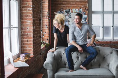 Yakışıklı bir adamla açık renk saçlı bir kadının, ortasında tuğla duvarlar ve kanepe olan bir odada çekilmiş bir fotoğrafı..