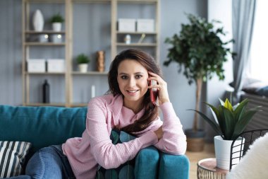 Çekici genç bir kadın cep telefonuyla konuşuyor ve evdeki kanepede otururken gülümsüyor. Akıllı telefondan ara..