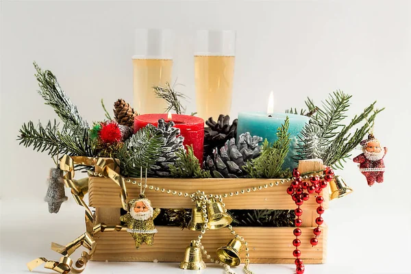 wooden box full of winter ornaments, pine leaves, cones and colorful candles on white background with two glasses of champagne