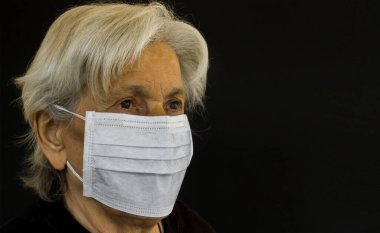 ISTANBUL, TURKEY - 24 Mart 2020: 65 yaşın üzerinde kadın siyah arkaplanda fotokopi alanı ile korunmak için hijyenik yüz maskesi kullanıyor.