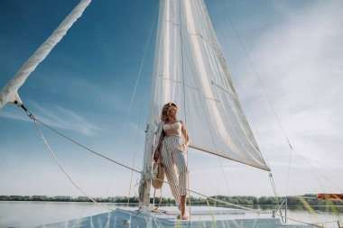 Beyaz yelkenli bir yatta duran kadın gün batımını seyrediyor. Yatta tatilde olan bir model.
