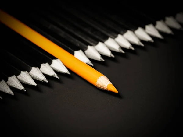 Black and white picture of the pencils with one orange coloured pecncil in the middle on a black backgound. Not like the others, being different.